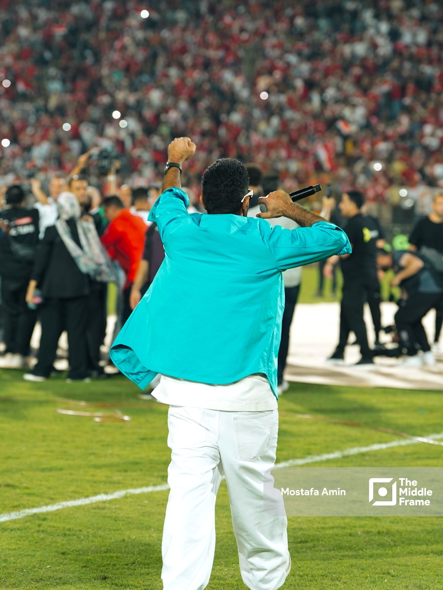 A man in a blue jacket and white pants is speaking to the crowd of a football stadium