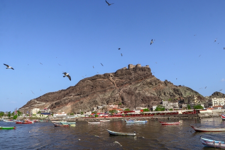 A beautiful scene of a volcanic island with the birds flying under a clear sky and small fishing boats anchored in the harbor