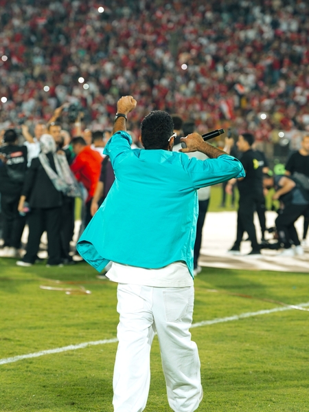 A man in a blue jacket and white pants is speaking to the crowd of a football stadium